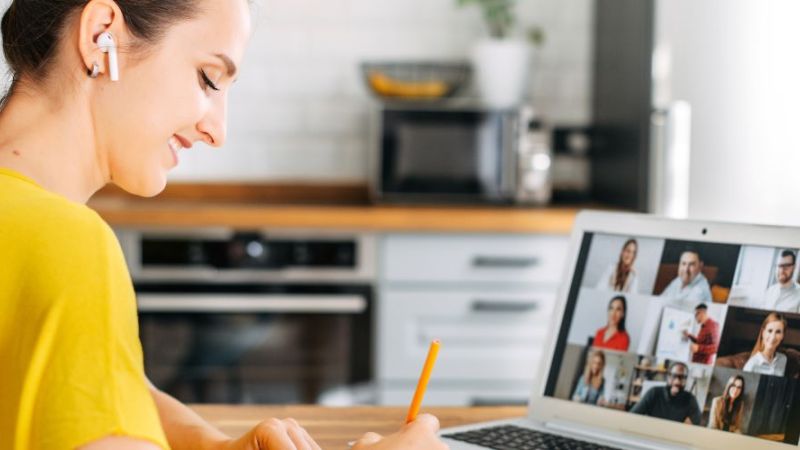 Eine Frau sitzt vor einem Laptop, nimmt an einer Videokonferenz teil und macht sich dabei Notizen.