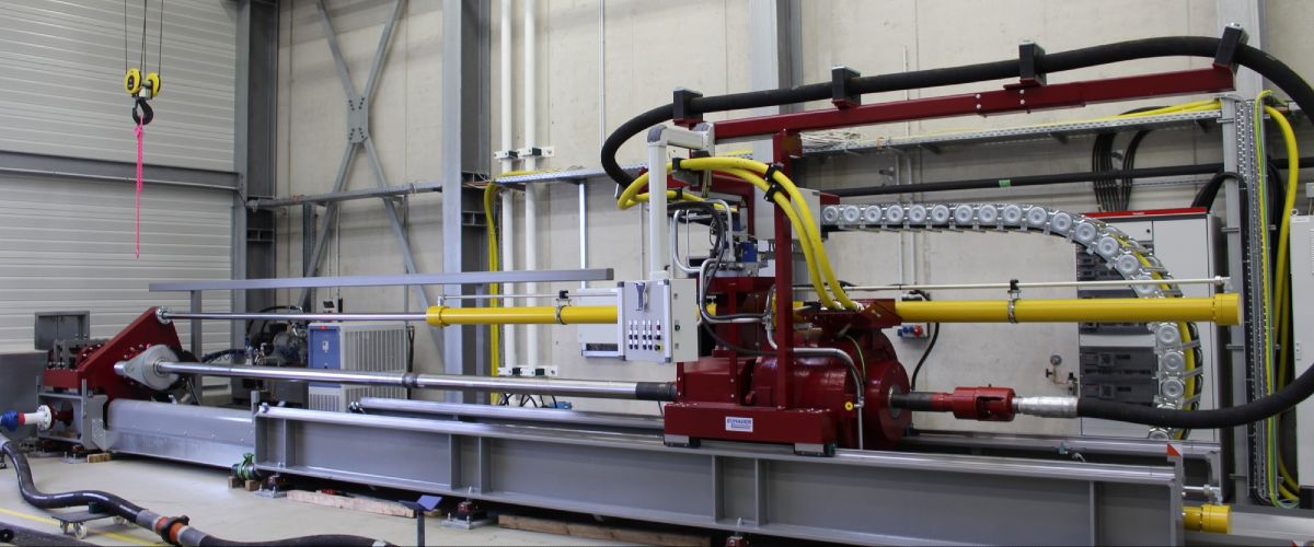 Output configuration of the test rig. The tests are being carried out with a horizontally mounted drilling rig. The drive (red, in the middle of the picture) drives the drill pipe, which protrudes from the hall onto the test site.