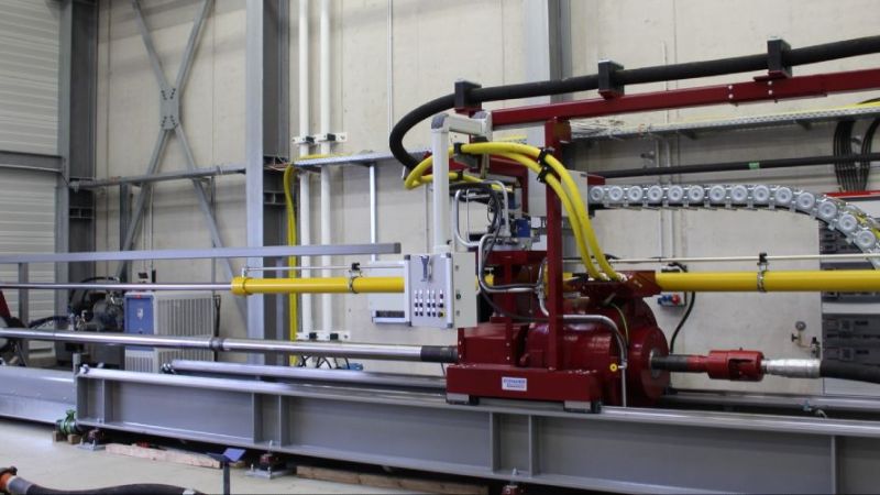 Output configuration of the test rig. The tests are being carried out with a horizontally mounted drilling rig. The drive (red, in the middle of the picture) drives the drill pipe, which protrudes from the hall onto the test site.