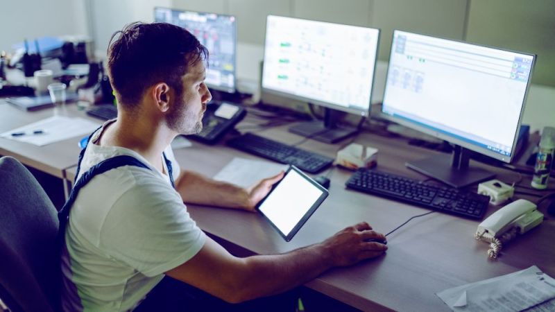 A worker sits in front of a computer and checks measured values with a tablet in his hand.