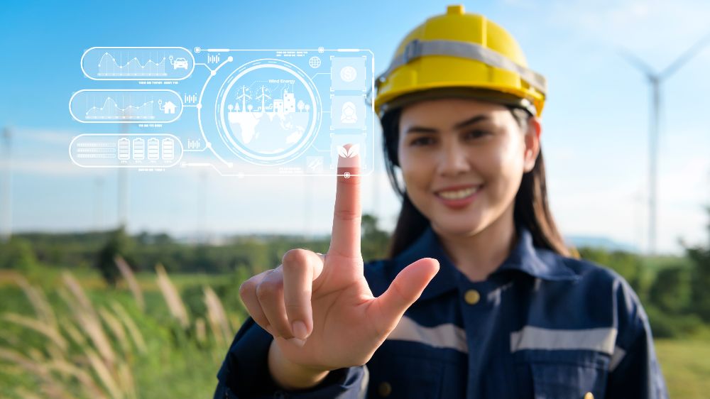 A woman wearing a yellow helmet stands in a field in front of a wind turbine.